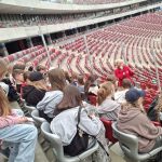 Uczniowie siedzą na krzesełkach na Stadionie Narodowym - słuchają Pani przewodnik